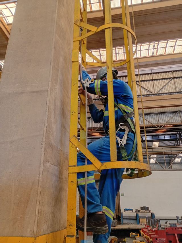 Trabalhador com cinto e talabarte sobe uma escada tipo marinheiro com guarda-corpo.