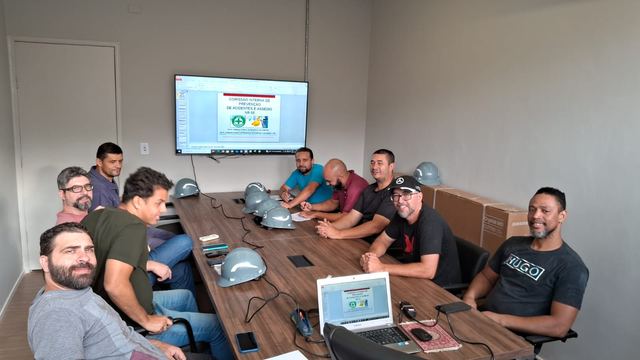 Turma reunida em volta de uma mesa, com capacetes e uma apresentação sobre a CIPA na tela.