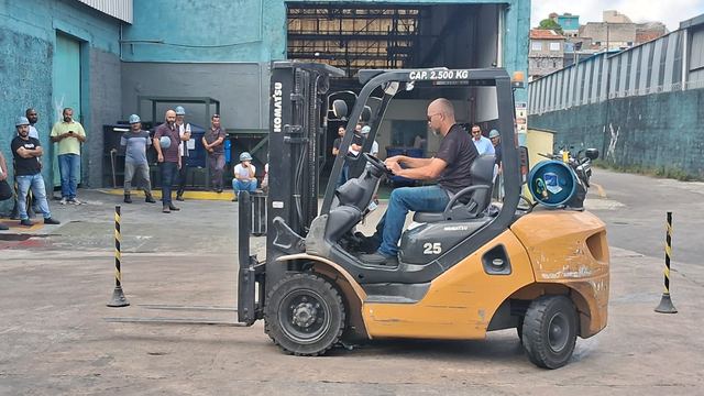 Operador conduz uma empilhadeira entre cones; ao fundo, a turma acompanha.