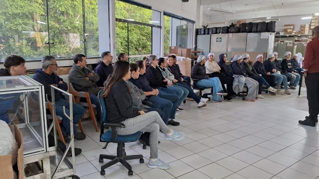 Plateia sentada em semicírculo assiste a uma palestra em um galpão.
