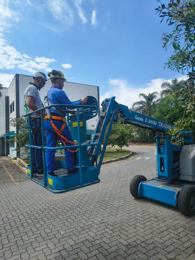Dois trabalhadores com cinto de segurança no cesto de uma plataforma elevatória, ao ar livre.