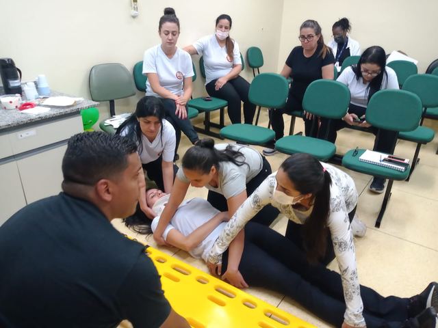 Participantes praticam o atendimento a uma pessoa deitada sobre uma prancha, em sala de treinamento.