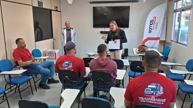 Instrutora fala a motoristas de camiseta vermelha sentados em uma sala de treinamento.